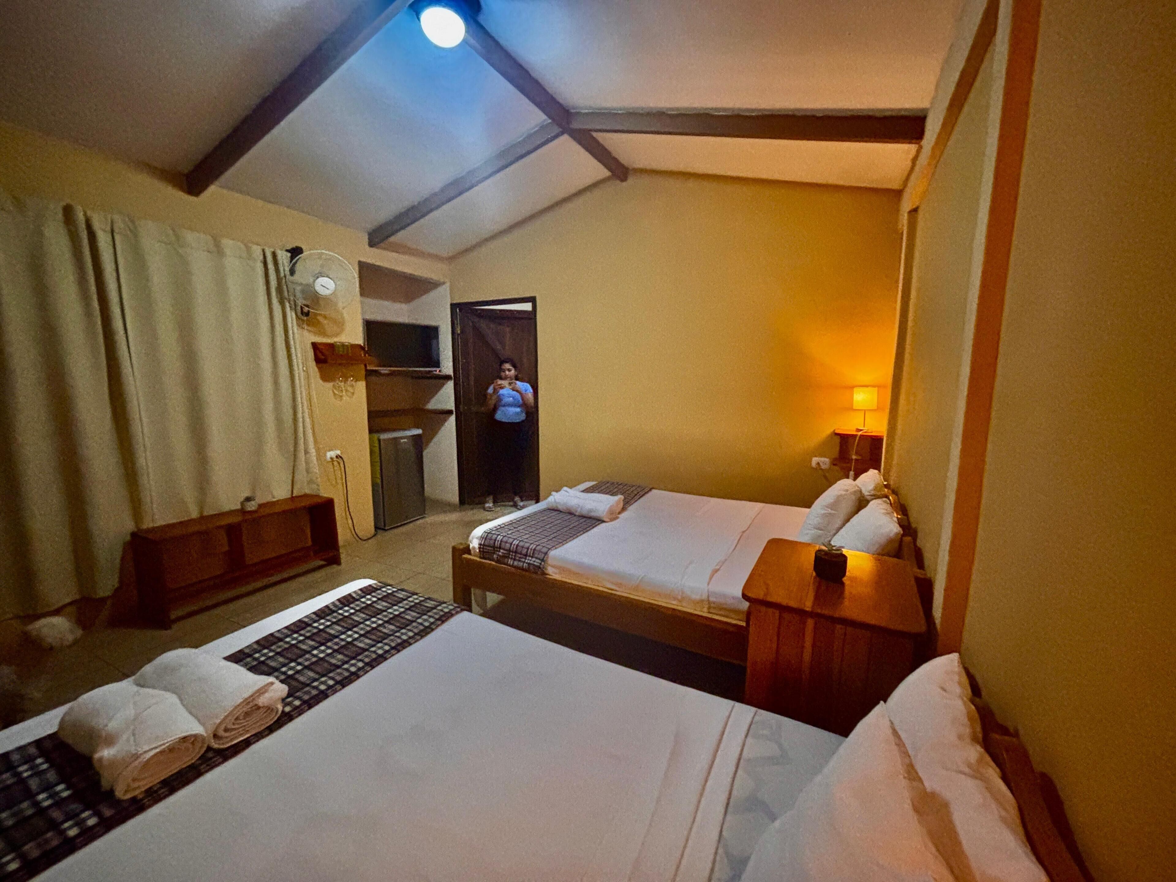 Habitación de hotel con dos camas, un escritorio, una silla, una mesita, un espejo, un ventilador y una puerta que da al baño.