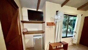 Une pièce de taille modeste, équipée d’un téléviseur posé sur une étagère, d’un petit réfrigérateur, d’un ventilateur et d’une fenêtre donnant sur un paysage verdoyant.