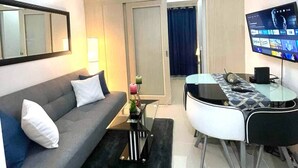 A modern living room with a grey sofa, a coffee table, a TV, and a dining area with a glass table and chairs. - Jazz Residences Tower B +Wifi & Netflix (Makati)