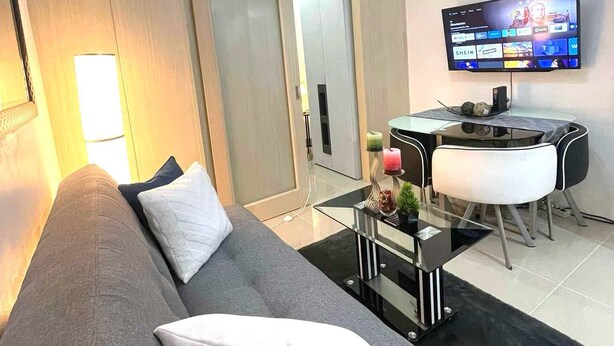 A modern living room with a grey sofa, a dining table with chairs, a flat-screen TV mounted on the wall, and a chandelier. - Jazz Residences Tower B +Wifi & Netflix (Makati)