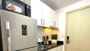 A compact kitchen with a refrigerator, a microwave, and a sink. There is a small countertop with a knife block and a plant. The cabinets are white, and the floor is tiled. - Jazz Residences Tower B +Wifi & Netflix (Makati)