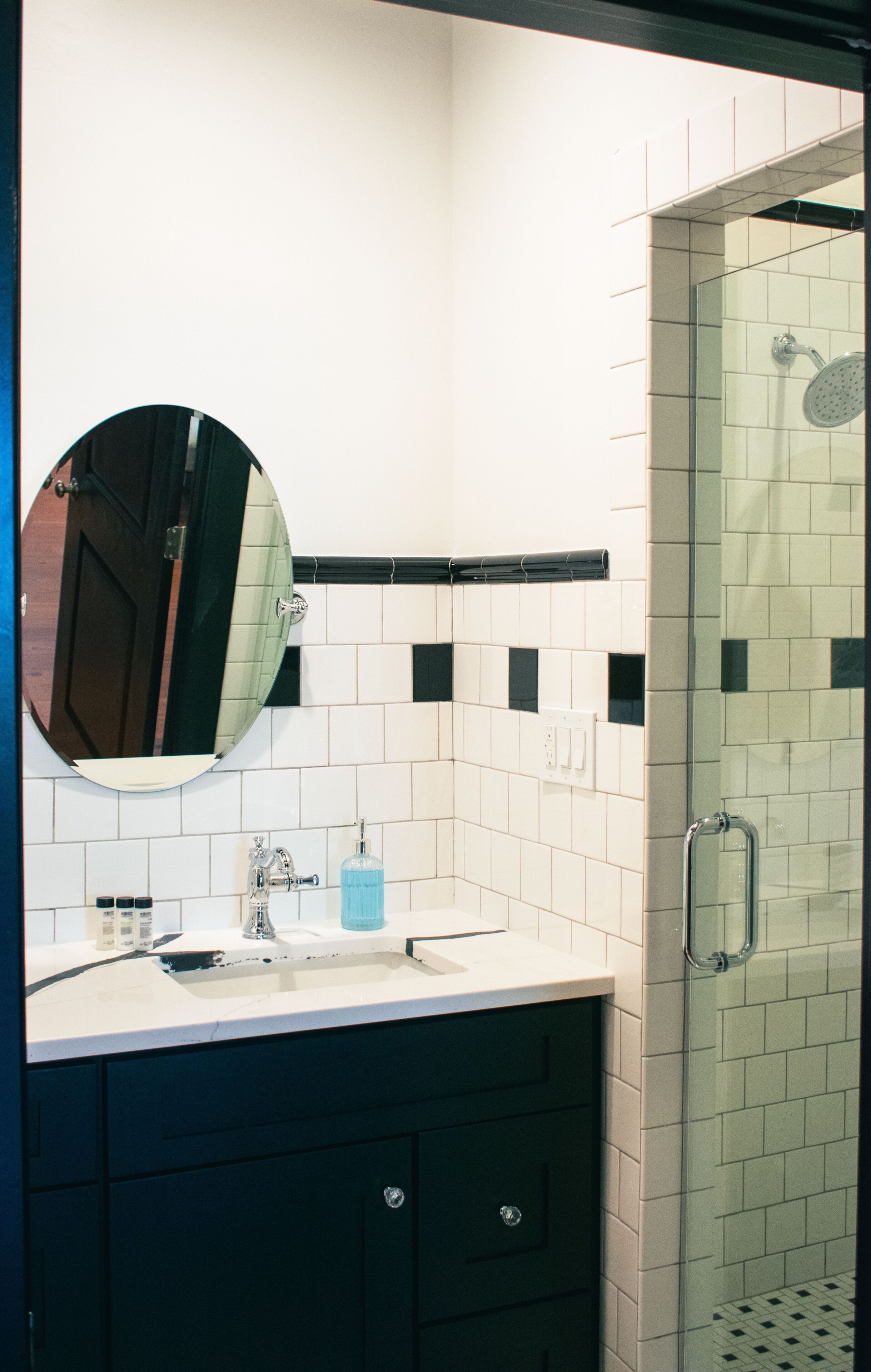 A bathroom with a white tiled wall, a round mirror, a sink, a glass shower enclosure, and dark-colored cabinets.