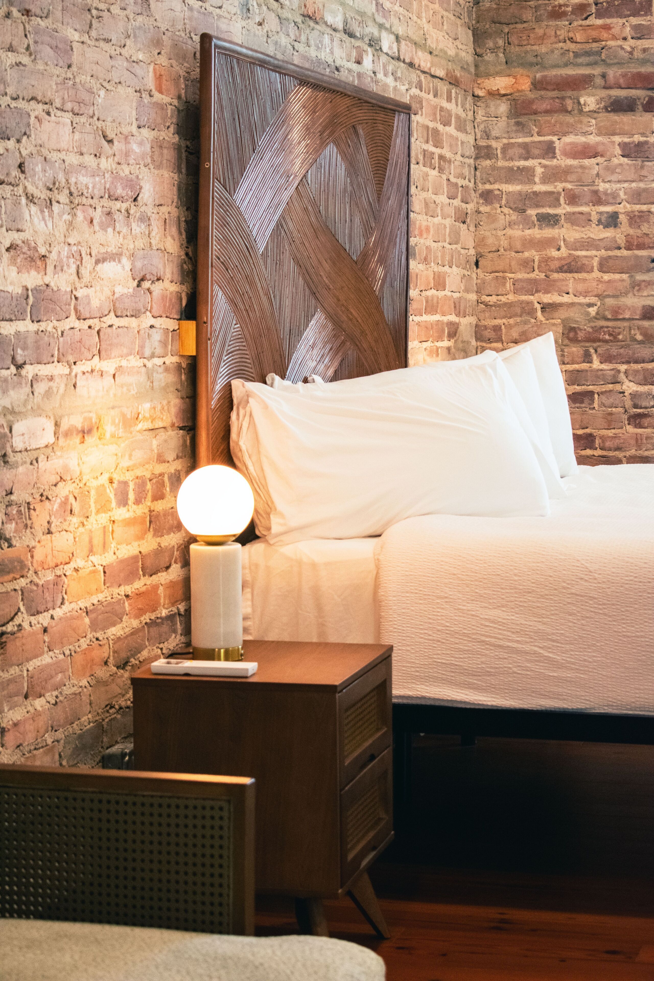 A bedroom with a brick wall, a wooden headboard, a bedside table with a lamp, and a bed with white bedding.