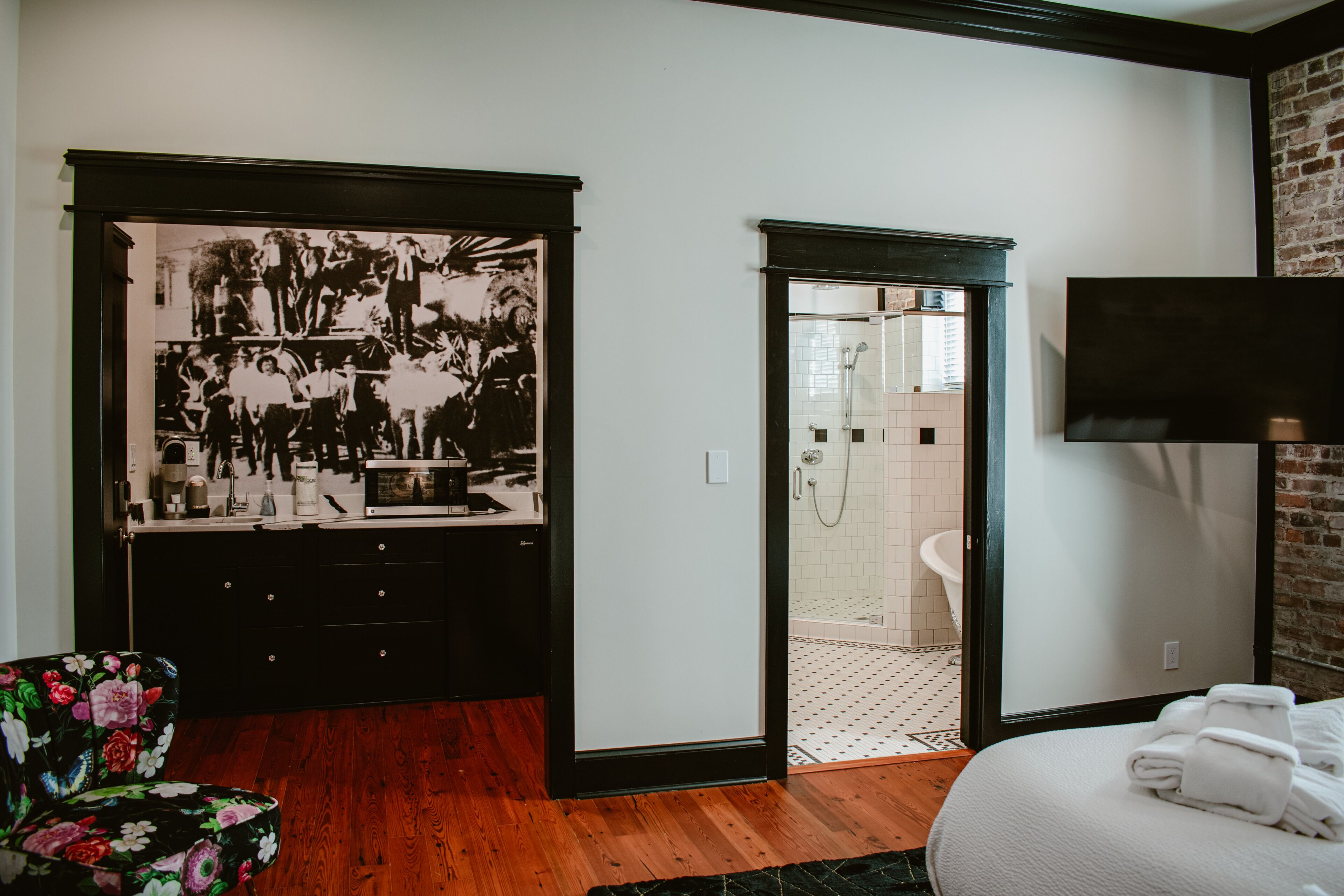 Una habitación con piso de madera, un espejo de marco negro, una cama blanca, un armario de marco negro y una puerta que da a un baño.