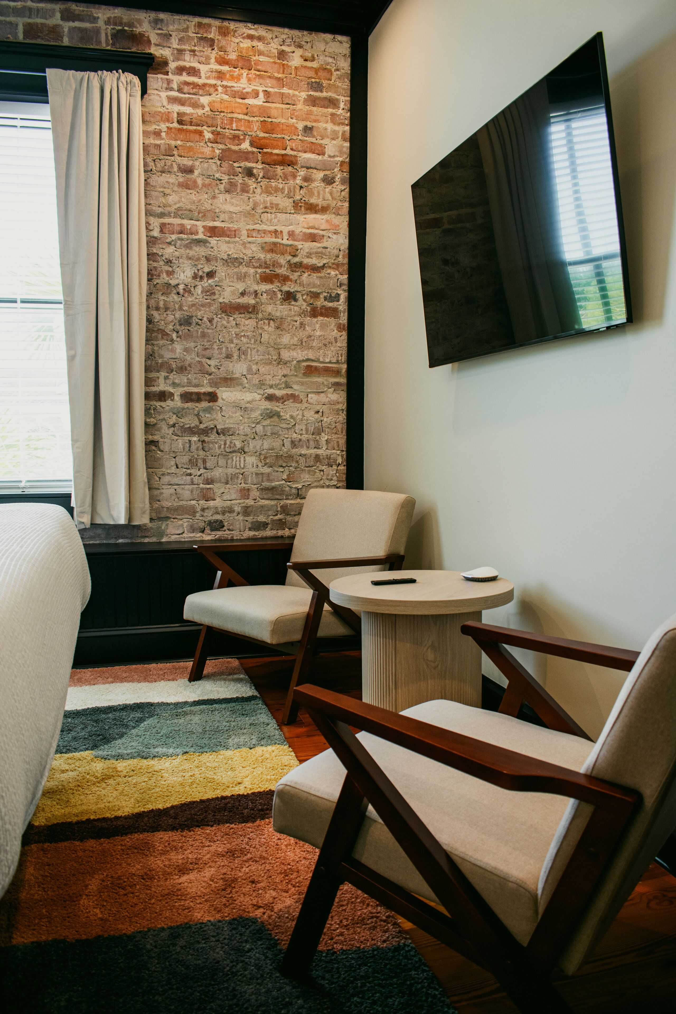 A hotel room with a brick wall, a bed, two chairs, a small round table, and a TV mounted on the wall.