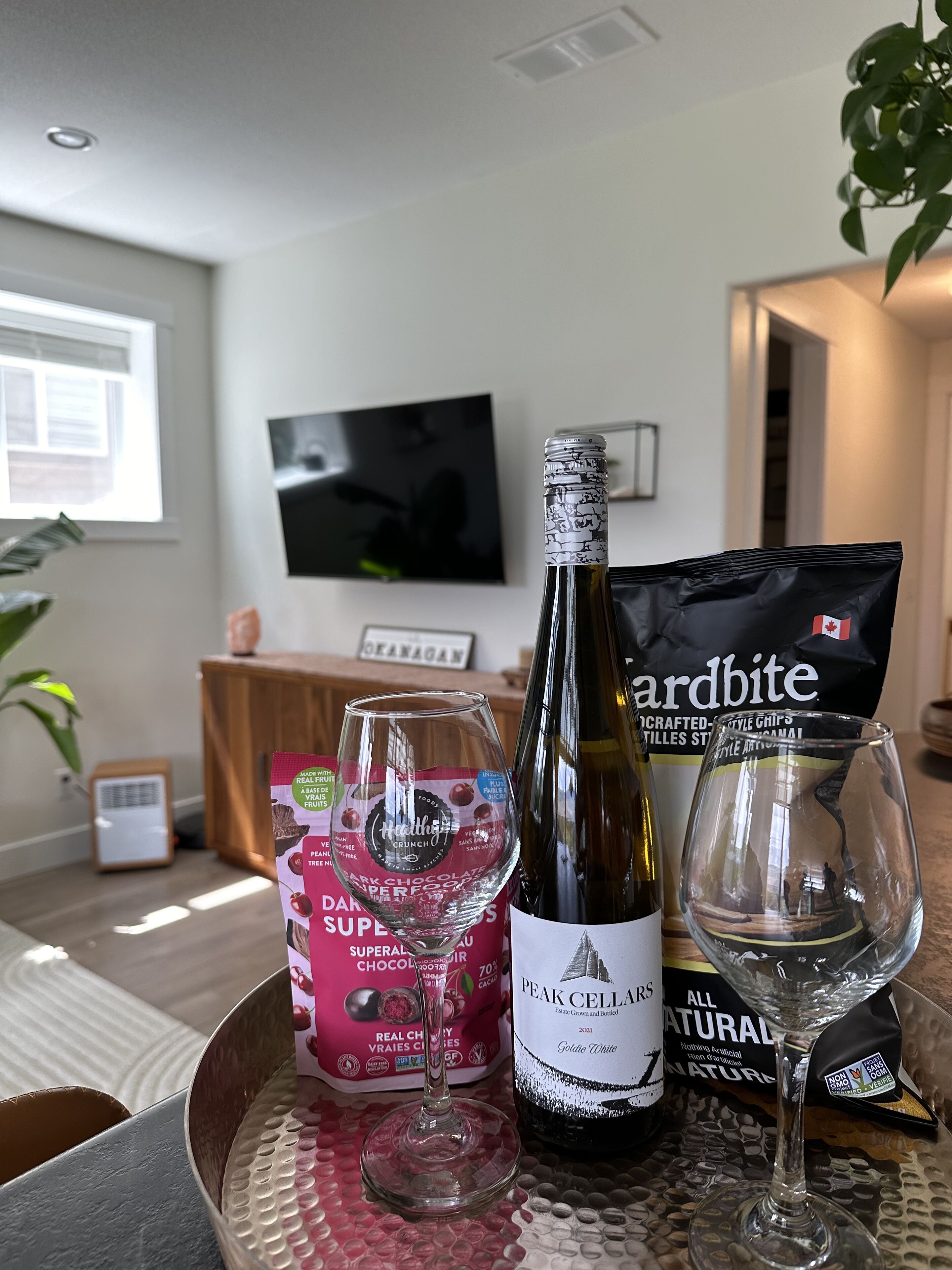 Ein Esstisch mit einer Weinflasche, einem Glas, einer Tüte Snacks und einer Schokoladenschachtel.