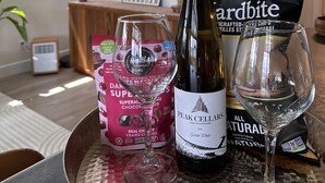 A dining table with a bottle of wine, a glass, a packet of snacks, and a box of chocolate.
