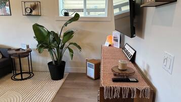 A living room with a large potted plant, a coffee table with a remote control, a framed welcome sign, and a TV mounted on the wall.