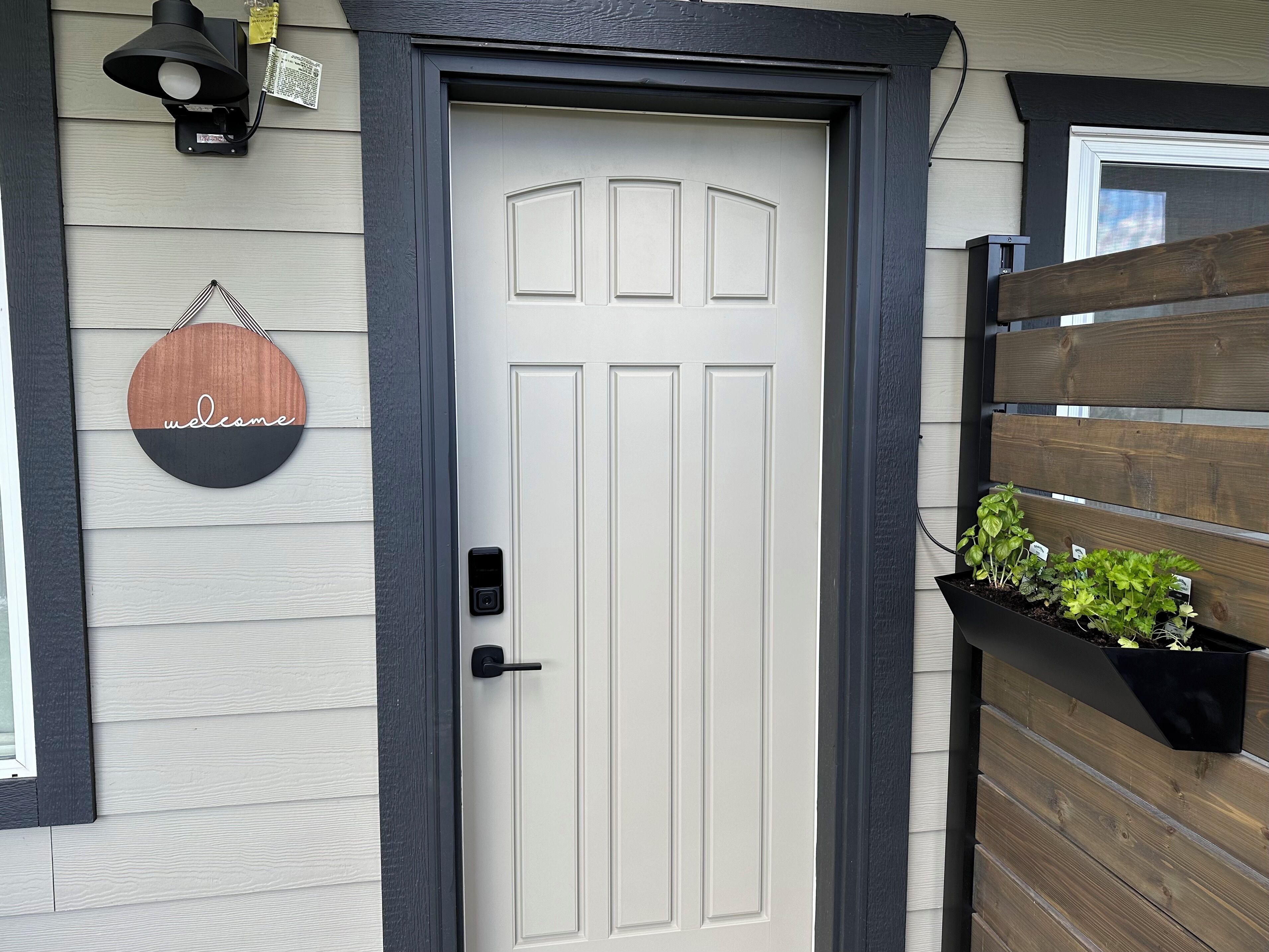A white door with a black handle, a welcome sign, a security camera, and a planter with plants.