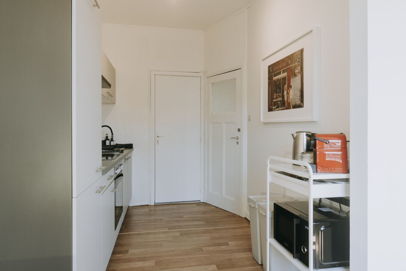 A narrow hallway leading to a white door, with a kitchen area on the left and a microwave and toaster on a shelf to the right.