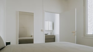 A minimalist bedroom with a bed, a bathroom with a mirror and sink, and a window with a brick wall view. - 1 bedroom apartment in Liege center, Place du Marché - 3rd floor (Liège)