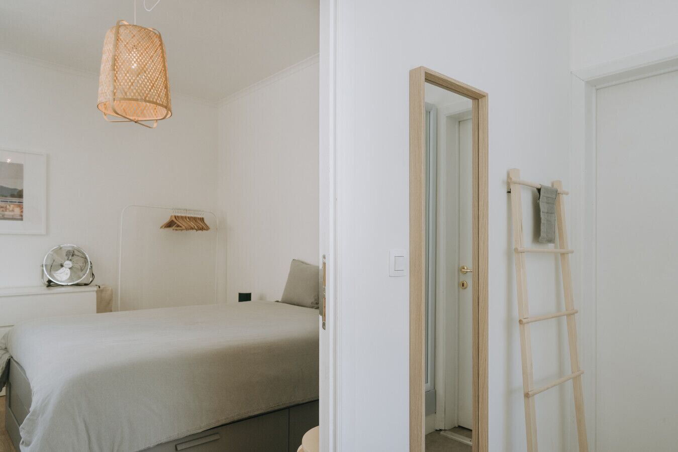 Een slaapkamer met een bed, een nachtkastje met een ventilator, een hanglamp en een deur die leidt naar een andere kamer met een houten ladder.