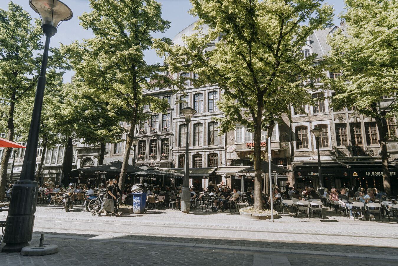 En brostensbelagt plads med udendørs siddepladser, historiske bygninger og træer.