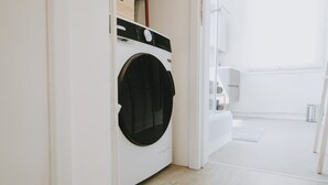 A modern laundry room with a front-loading washing machine, a wooden shelf, and a white tiled floor. - 1 bedroom apartment in Liege center, Place du Marché - 3rd floor (Liège)