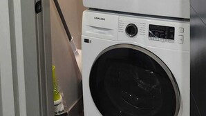 Un lave-linge et un sèche-linge Samsung à chargement frontal, superposés dans une buanderie, avec un plancher en bois et une porte donnant sur une autre pièce.