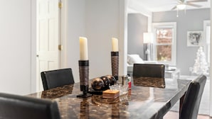 Una mesa de comedor con velas, saleros y pimenteros, y un objeto decorativo. Cuatro sillas rodean la mesa. En el fondo, se ve un ventilador de techo y una puerta.