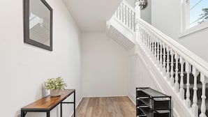 Un pasillo con suelo de madera, pared blanca, espejo con marco negro, una pequeña mesa con una planta y una escalera blanca con barandilla.