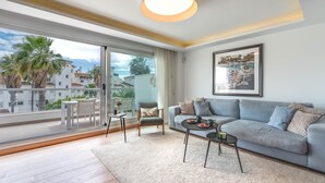 A modern living room with a grey sofa, a coffee table, and a chair. There is a large window with a view of palm trees and buildings.