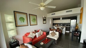 A modern living room with a red sofa, a grey armchair, and a coffee table. There is a ceiling fan, a kitchen in the background, and framed artwork on the wall. - Apartamento Vista Mare 16-301 (Samaná)