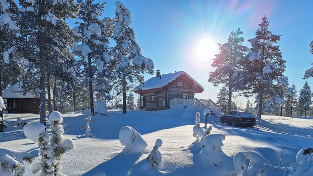 A log cabin with a snow-covered roof, surrounded by snow-laden trees and a clear sky.