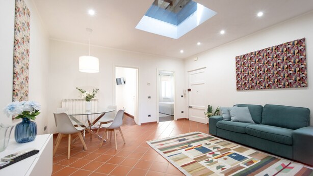 A modern living room with a green sofa, a patterned rug, a dining area with a glass table and chairs, and a skylight above.