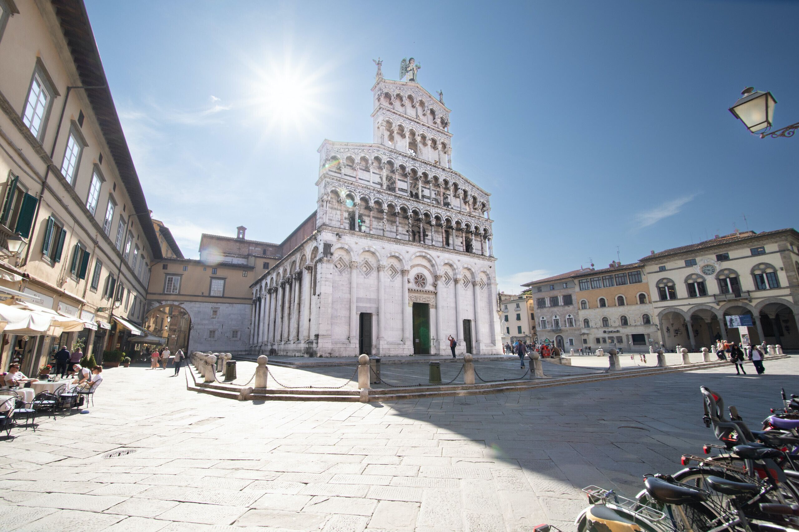 A spacious square with a prominent historical building featuring multiple arches and a statue on top.