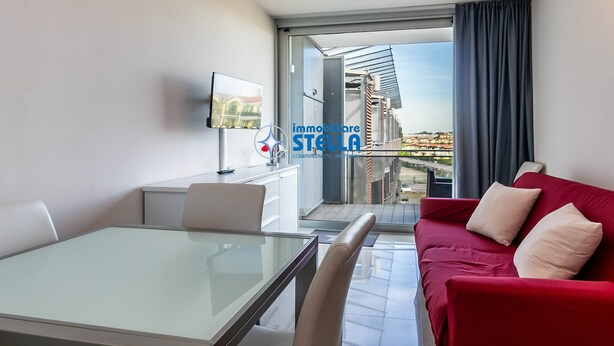 Une chambre d’hôtel moderne avec une table à manger en verre, un canapé rouge, un téléviseur fixé au mur et un balcon offrant une vue sur la ville.