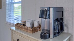 Een koffiezetapparaat op een aanrecht, met een mand mokken en een raam met zonwering.