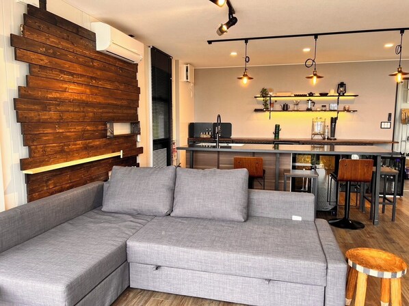 A modern living room with a grey sectional sofa, wooden wall paneling, a kitchen area with a bar, and pendant lighting.