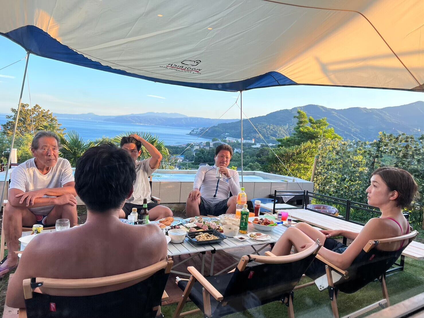 A group of people dining under a tent with a scenic view of the sea and hills.