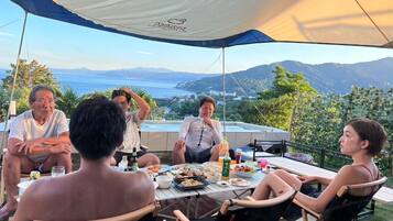 A group of people dining under a tent with a scenic view of the sea and hills.