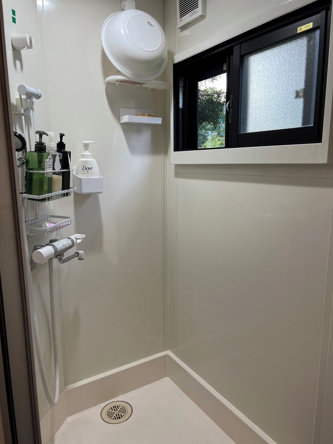 A small, enclosed shower area with a window, a hand dryer, and toiletries on a shelf.