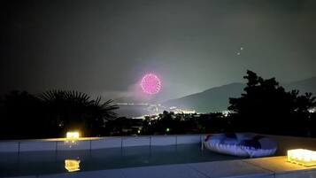 A nighttime view of a pool area with a distant fireworks display, a mountain in the background, and a clear sky.