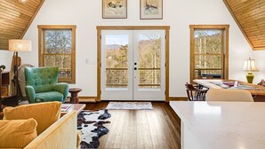 A spacious living area with a high ceiling, wooden beams, a large green armchair, a dining table, and a view of trees through the glass doors. - Modern A-Frame Away from it All! (Marshall)