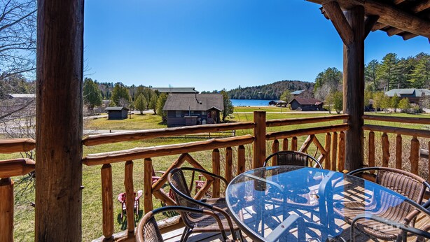 A wooden deck with a glass table and wicker chairs, overlooking a lake and buildings.