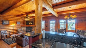 A log cabin interior with a stone fireplace, wooden furniture, a kitchen with a sink, and a large window overlooking the outdoors.