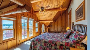 A cozy wooden cabin bedroom with a large bed, a ceiling fan, and a view of the outdoors through large windows.