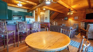 A log cabin interior with a wooden dining table, chairs, a fireplace, and a kitchen area.