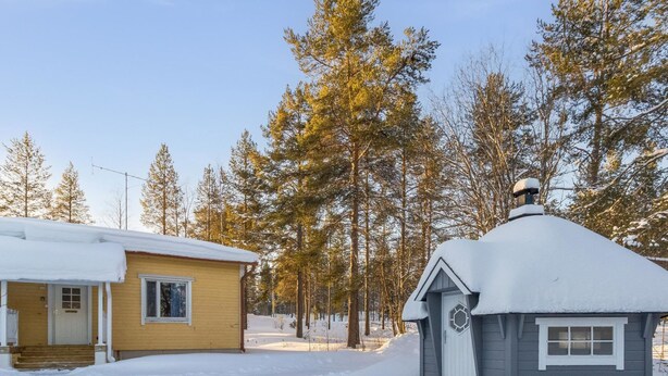 A cabin with a snow-covered roof, a wooden porch, and a small attached shed with a chimney.