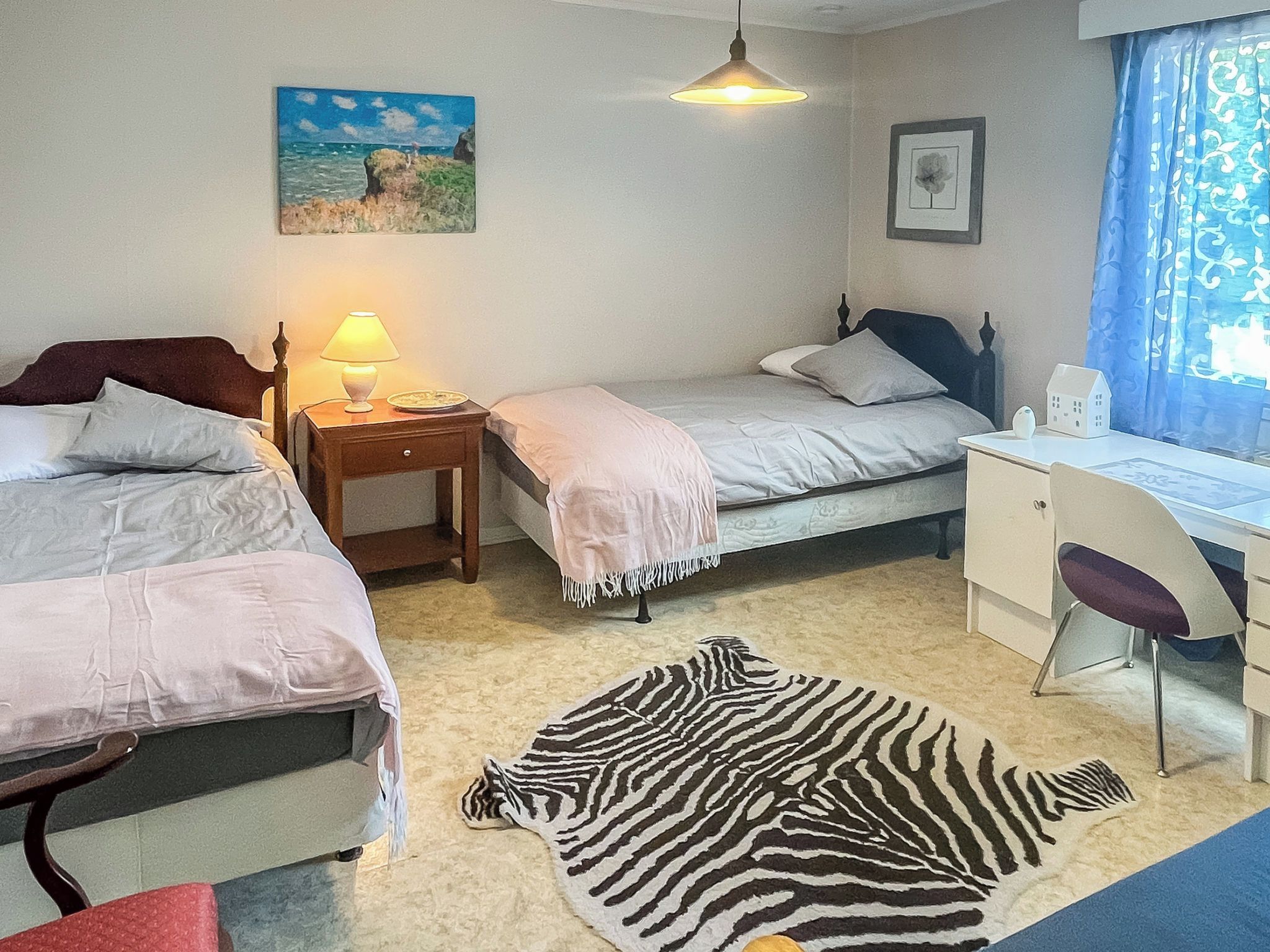 Une chambre avec deux lits, un bureau, une chaise, une lampe, un tapis à motifs zébrés et des œuvres d’art accrochées aux murs.