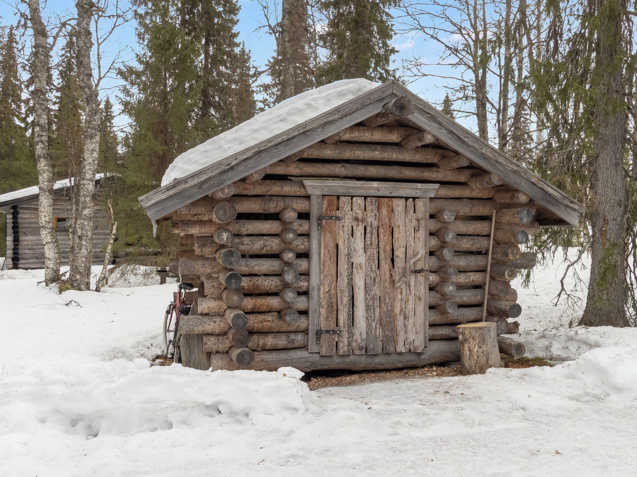 A rustic log cabin with a weathered wooden door, surrounded by snow and trees.