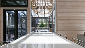 Een moderne eetruimte met een grote witte tafel, metalen stoelen en een houten plafondlamp.
