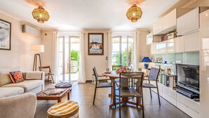 Ein modernes Wohnzimmer mit Sofa, Couchtisch und Hocker, ein Essbereich mit Stühlen und Tisch sowie eine Küche mit Schränken und Fernseher.