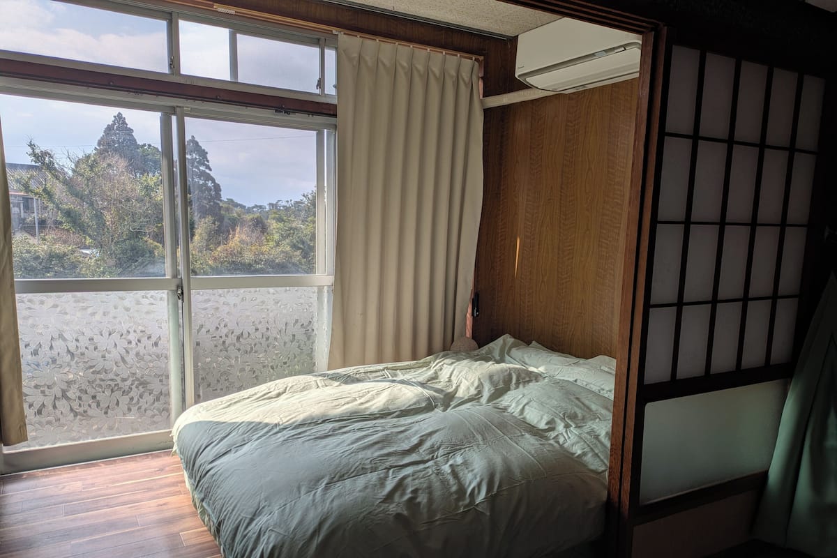 Une pièce avec un lit, une fenêtre avec des rideaux, une vue sur les arbres et une cloison en bois.