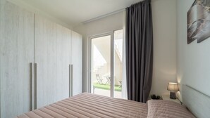 Un dormitorio con cama, un armario, una ventana con cortinas y una pequeña mesa con lámpara.