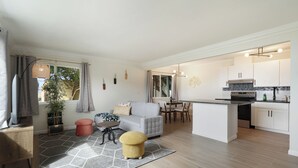Ein modernes Wohnzimmer mit einem grauen Sofa, einem kleinen runden Tisch und einem gelben Hocker. Es gibt eine Küchenzeile mit weißen Schränken und einer schwarzen Arbeitsplatte.