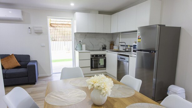 Une cuisine moderne avec des meubles blancs, un réfrigérateur en acier inoxydable et un coin repas comprenant une table en bois et des chaises blanches.