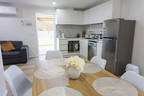 A modern kitchen with white cabinets, a stainless steel refrigerator, and a dining area with a wooden table and white chairs. - Modern 2BR Guest House in Werribee, BBQ, Backyard & Prime Location (Werribee)