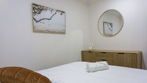 Une chambre à coucher avec un lit, une commode en bois, un miroir rond et un tableau représentant un arbre au bord de la mer.