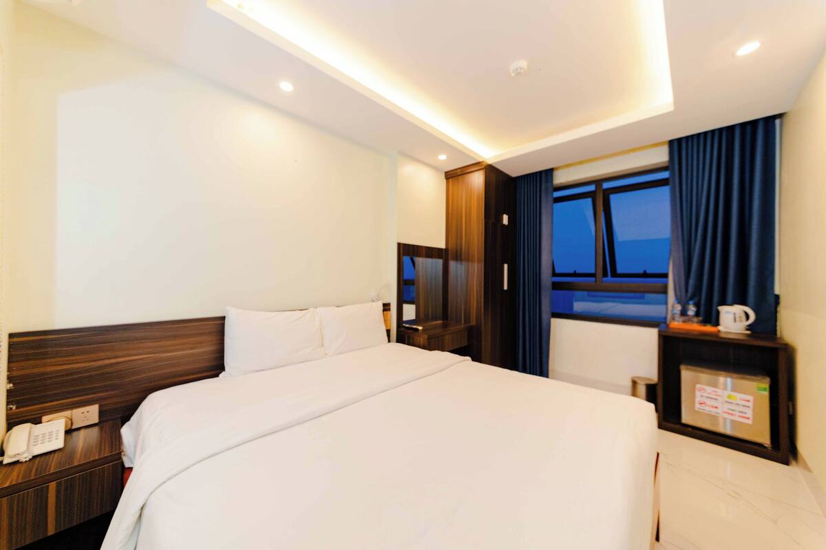 Ein Hotelzimmer mit einem großen Bett, einem Nachttischtelefon, einem Beistelltisch, einem Fenster mit Vorhängen und einem kleinen Tisch mit Tasse und Schild.